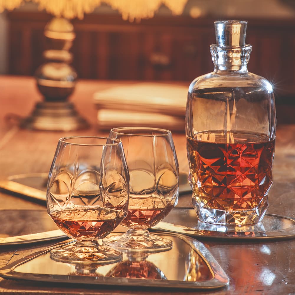 Гарафа за уиски или ликьори ALKEMIST RCR Italy от кристал Luxion, елегантна и модерна, италианско качество. Поставена върху маса осветена с приглушена светлина на нощно заведение в близост 2 чаши за коняк, поставени върху огледални подноси с налято уиски.