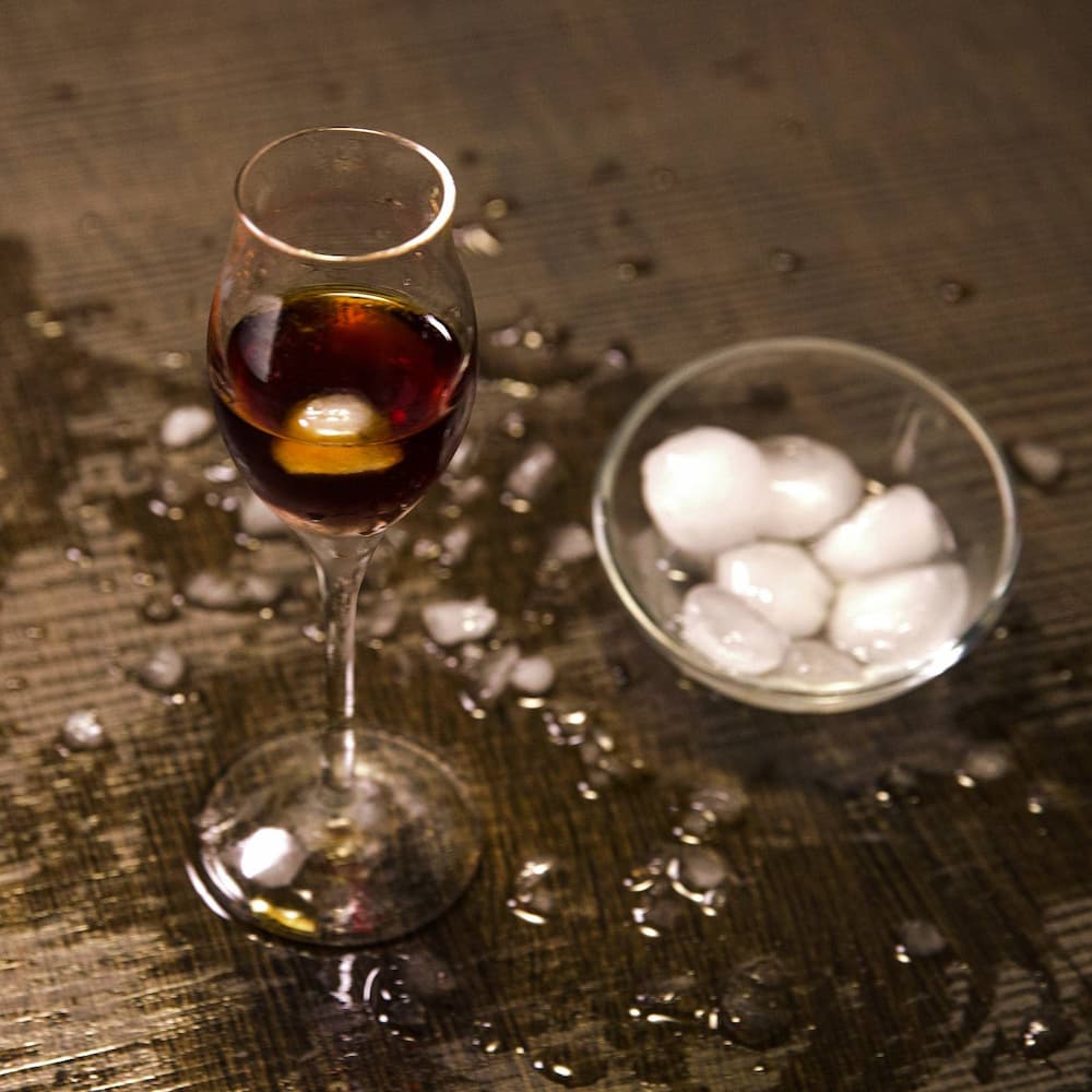 Eлегантни кристална чаша на столче invino с налята граппа и кубче лед, поставена върху дървен плот в бар обстановка с купичка с ледени кубчета.