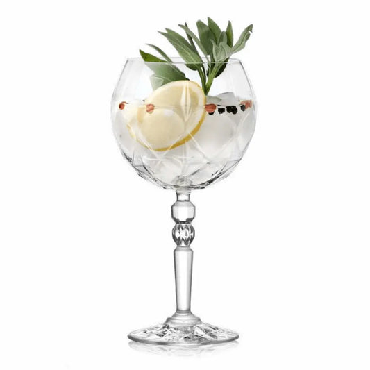 Елегантна кристална чаша за джин тоник ALKEMIST RCR Italy с декоративна основа, пълна с лед, лимон и зелени билки на бял фон.
