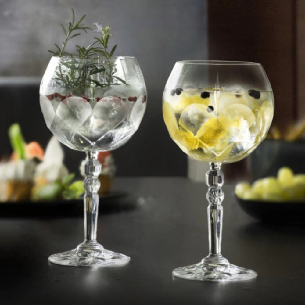 Комплект кристални чаши ALKEMIST RCR Italy за коктейли - една с розмарин и червени плодове, друга с лимон и подправки върху черна маса в елегантна бар обстановка с приглушена дневна светлина.
