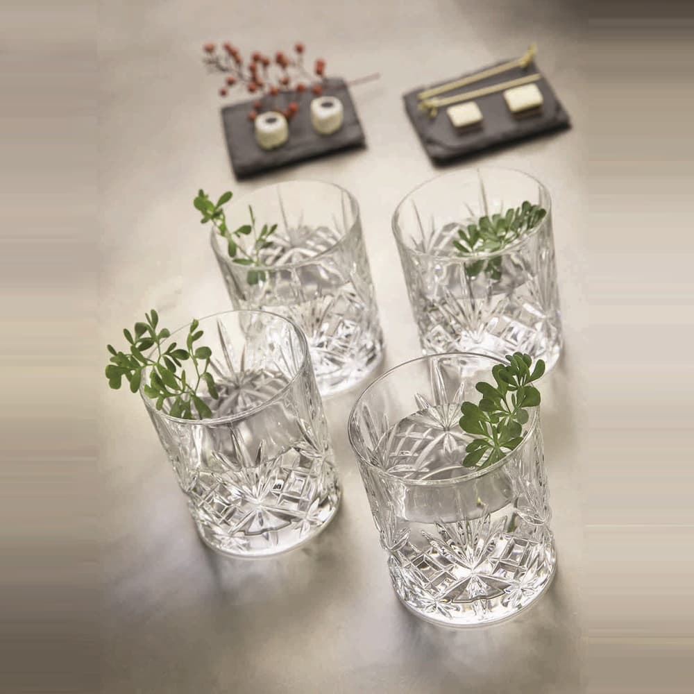 Кристални чаши MELODIA с класическо диамантен гравиран мотив, с налята вода и украсени с свежи зелени цветя в азиатски стил с 2 подноса с ордьоври на задния фон върху плот в цвят слонова кост.